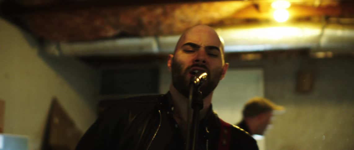 A bald man with a beard passionately sings into a microphone while playing guitar his face showing intense emotion
