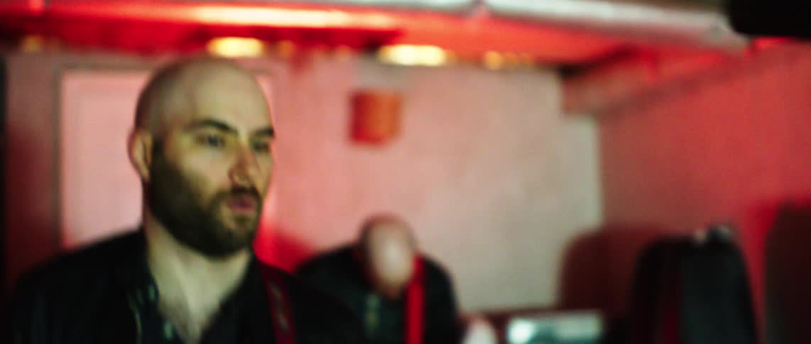 A bald man is singing and playing guitar while other man in the back is playing guitar in a studio setting under a red light