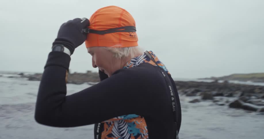 Older friends mostly wearing wet suits wade into the ocean water under an overcast sky