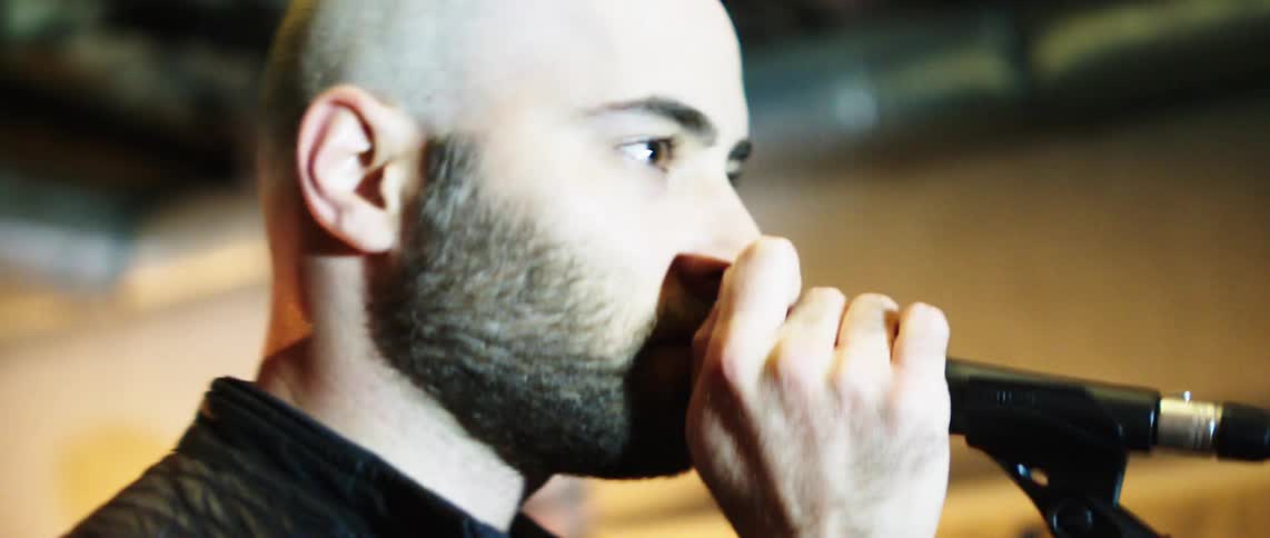 A bald man singing on a microphone on an indoor setting under a blinking light