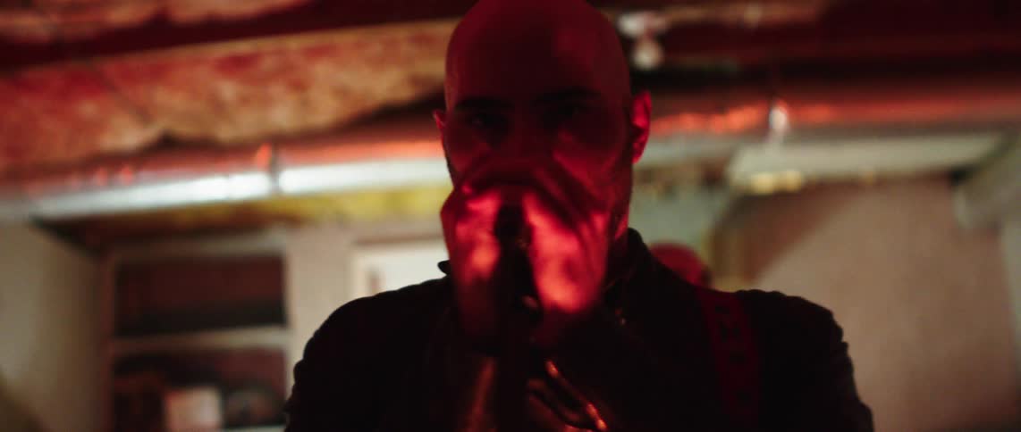 A bald man with a beard grips a microphone and stares intensely forward under dramatic pink lighting