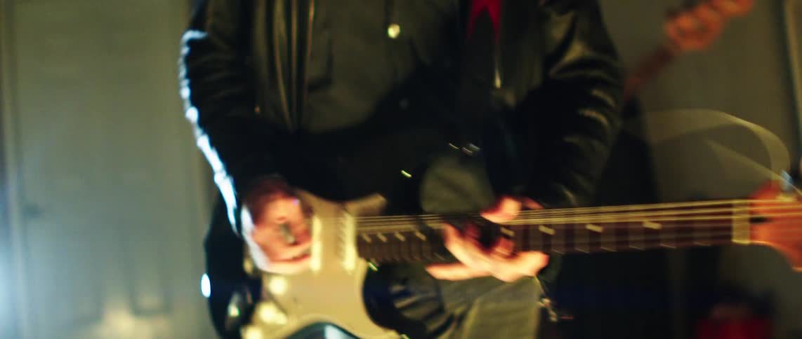 Guitarists play in a messy room lit by colored lighting