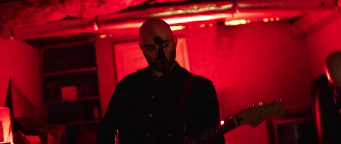 Guitarists perform in a messy room lit by colored lights
