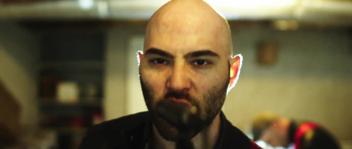 A bald man singing in a microphone in a studio setting with another man playing guitar in the background