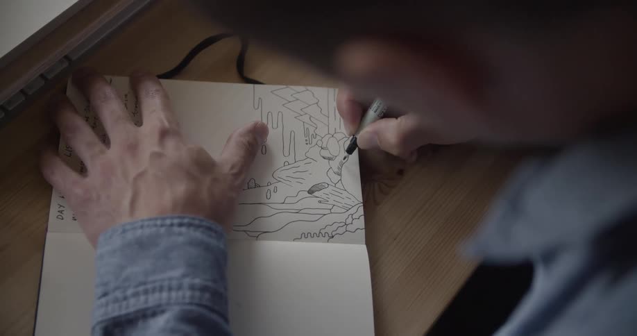 A person is holding a pen and drawing a sketch on a notebook that is on a wooden table