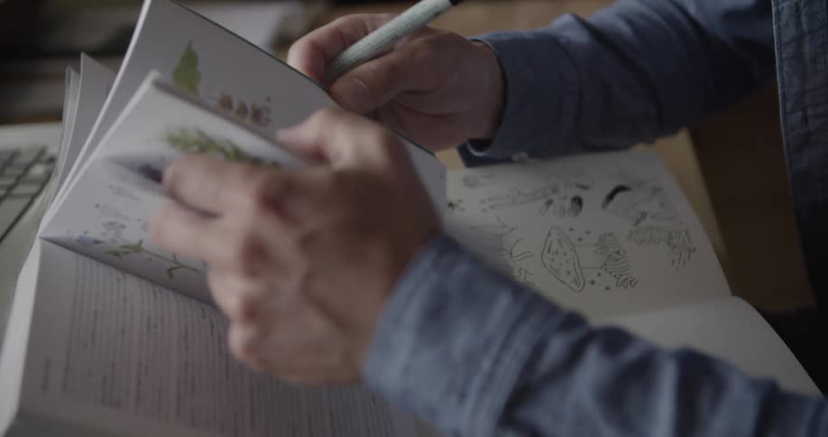 Pair of hands holding a pen leafing through a book with drawings
