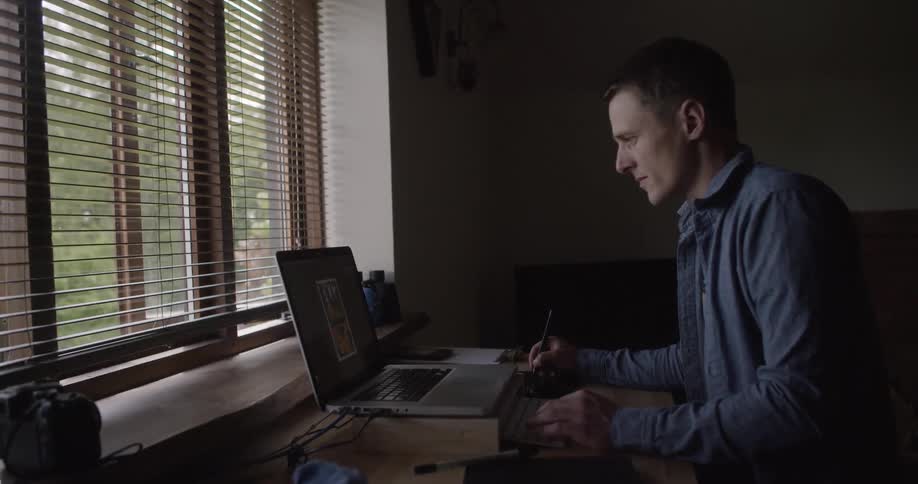 A man is working at a desk on his laptop using a stylus by the window