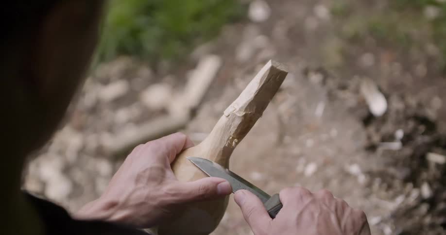 Man using a knife to sharpen a piece of wood in his hand while in the woods