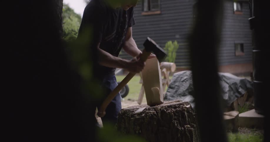 A man wearing a black shirt and jeans stands in a backyard using an axe to split a log on a large stump with a dark wooden building in the background