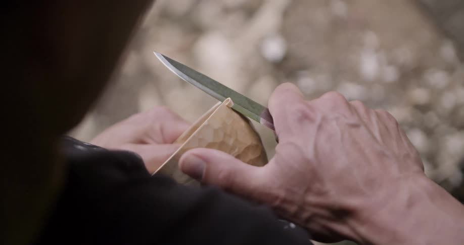 A persons hands holds a knife and a piece of wood carving wood with the knifes blade