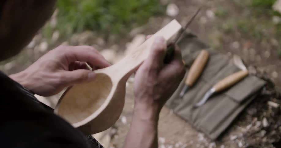 Man carving a wooden spoon while camping in the woods at midday
