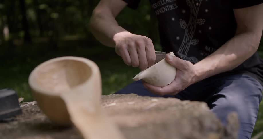 Man smoothing a freshly carved wooden spoon with sandpaper at midday in the woods sitting on a tree trunk with a larger finished spoon beside him