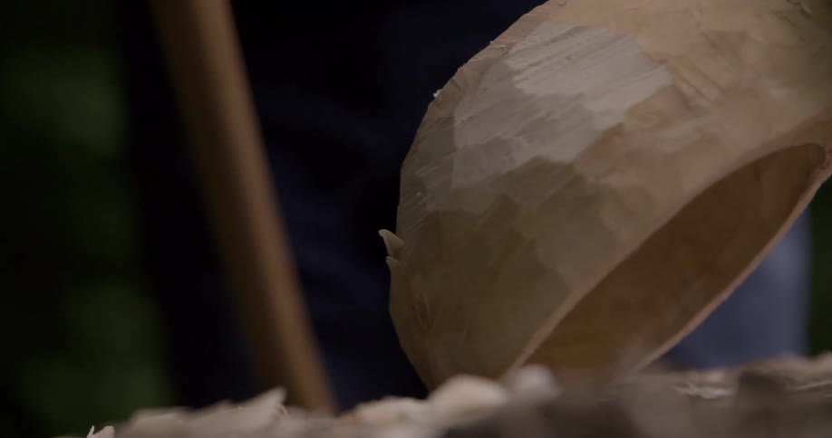 A persons hand uses a chisel to carve a piece of wood creating wood shavings