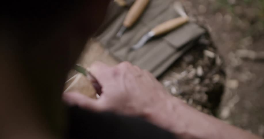 A person carves a wooden cup with a knife on a tree stump outdoors
