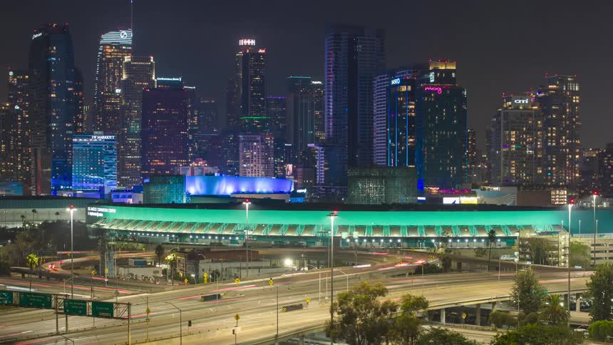 The video shows a timelapse of a highway at night with a busy Los Angeles Convention Center and skyline in the background the lights of passing cars create long streaking trails on the road below