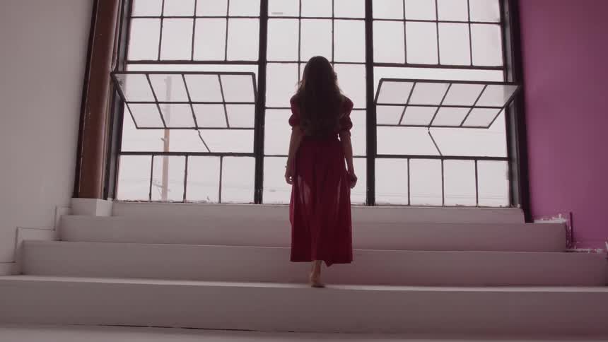 Woman with long brown hair wearing a long red dress walking up an indoor staircase barefoot at midday