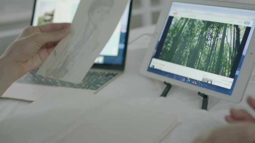 A woman holds up a sketch with a laptop and tablet nearby