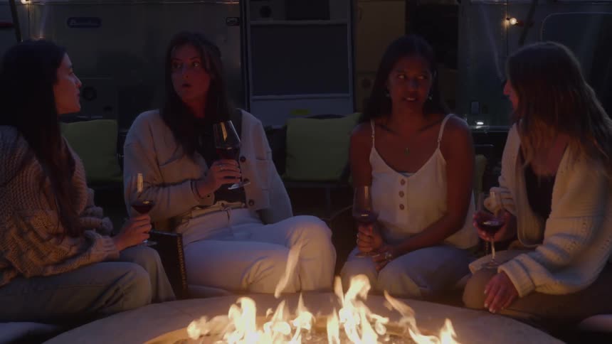 Four women sit around a fire pit drinking wine and talking