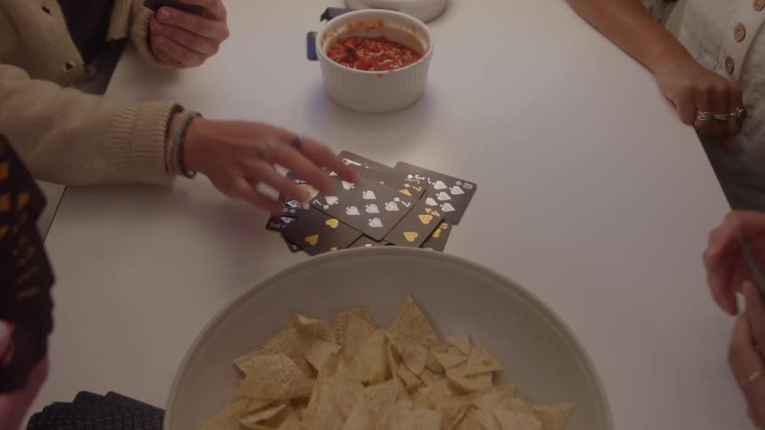 A highangle view shows a group of people sitting around a white table a large bowl of tortilla chips and a smaller bowl of red dip are in the center with several peoples hands visible as they reach for playing cards scattered on the table