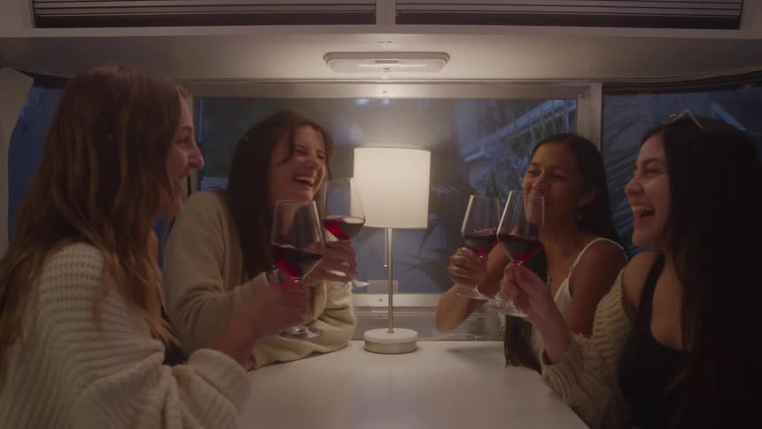 Four young female friends sitting at a table inside an rv holding wine glasses filled with wine clinking them together in a cheerful toast then drinking while laughing and smiling