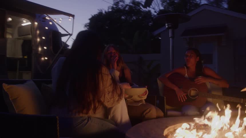 Group of friends sitting outdoors around a campfire talking and playing guitar