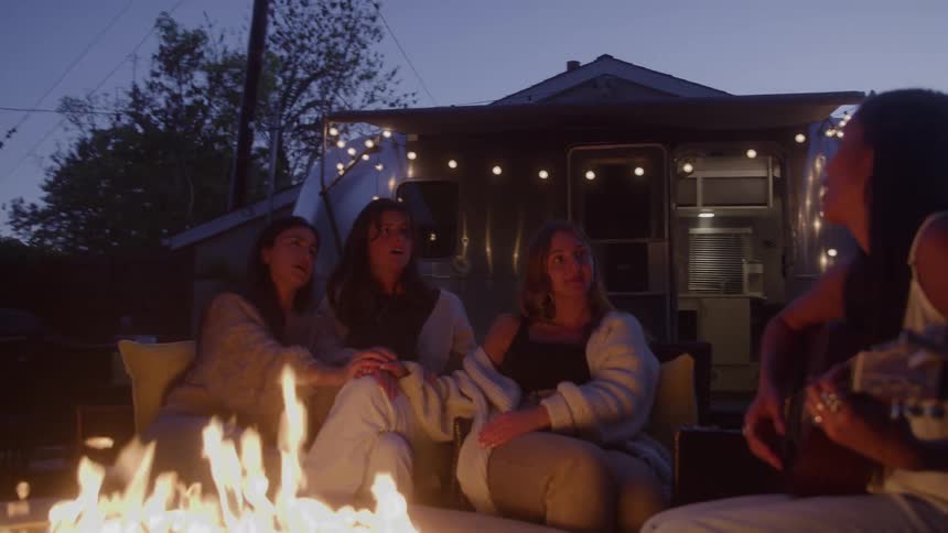 Four women sing and play guitar gathered around the fire pit