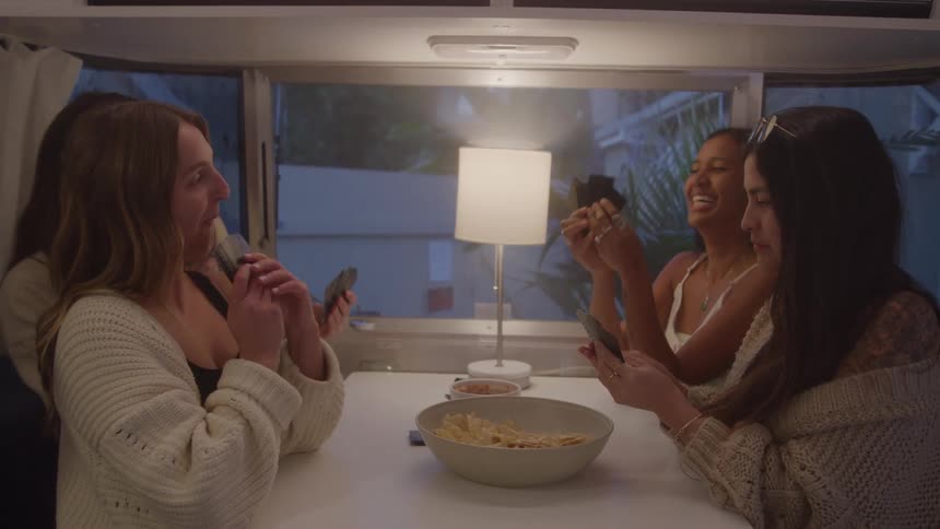 In this short video clip a group of four women are gathered around a table inside a camper they are all playing a card game together laughing and looking at their cards
