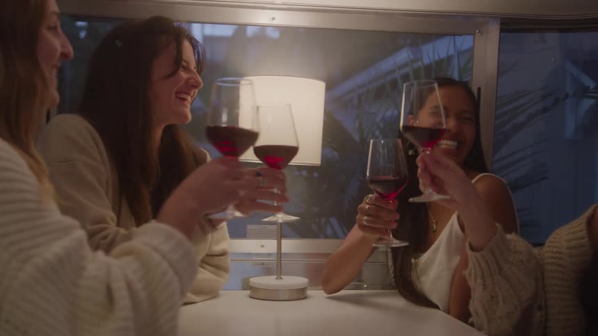 In this short clip four women are sitting together likely in a trailer or camper and toasting with glasses of red wine they all appear to be having a good time laughing and smiling as they clink their glasses together and take a sip
