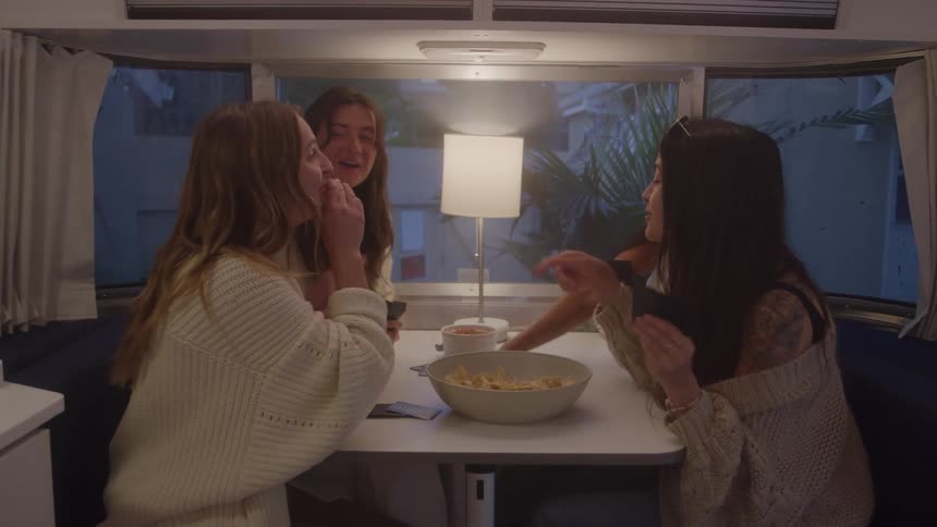Four women are gathered around a table inside a trailer playing a card game they are laughing and talking with one woman holding a fan of cards in her hand and a bowl of chips sitting on the table between them