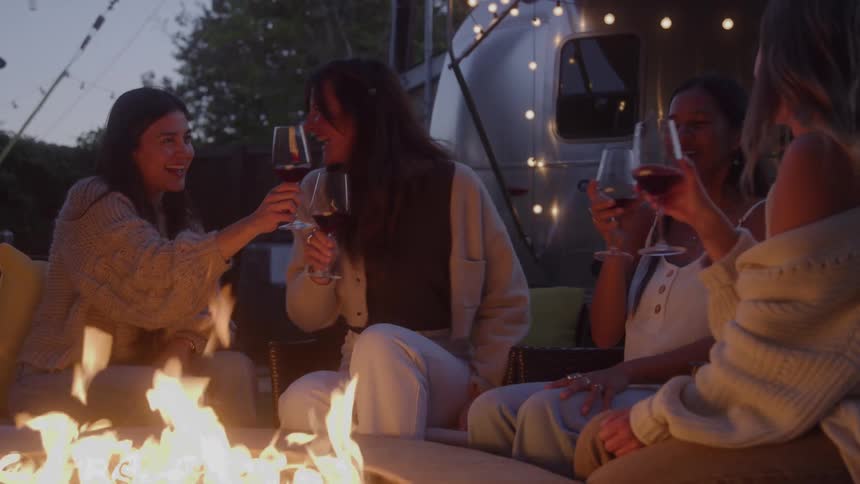 Four women are gathered around a campfire outside a silver camper van they toast with glasses of red wine smiling and laughing as they clink their glasses together and take a sip