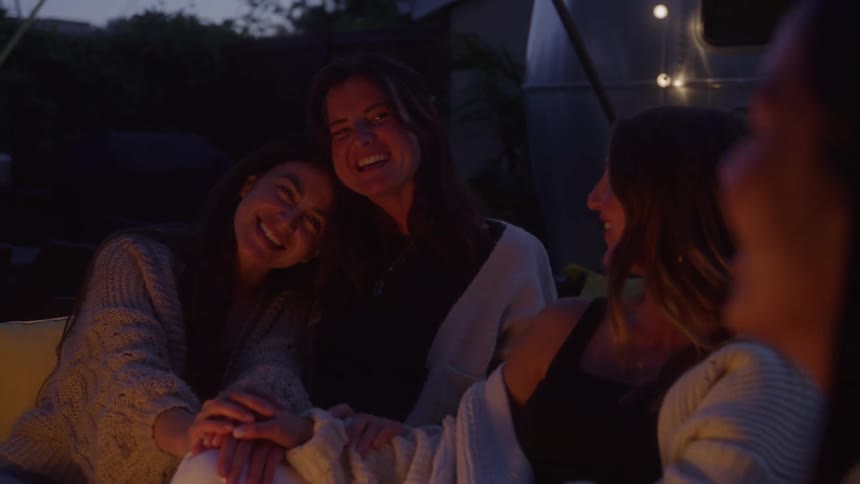 In this nighttime clip three women are sitting together outside in front of a trailer and what looks like a fire which casts a warm glow on their faces they are smiling and laughing looking happy and relaxed