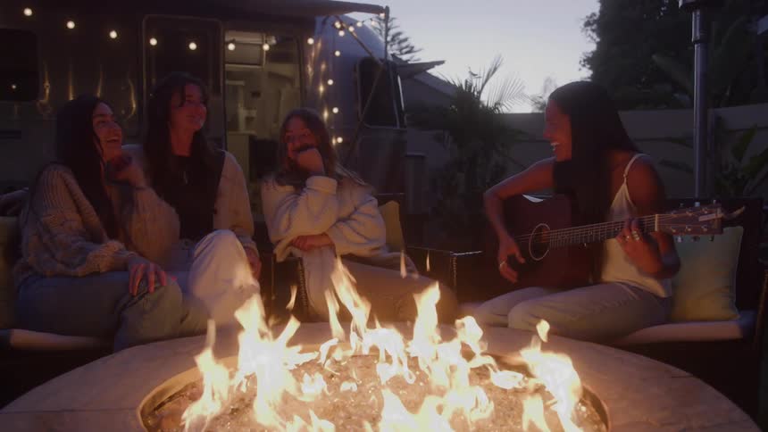 Three women watch while one plays guitar around the fire pit
