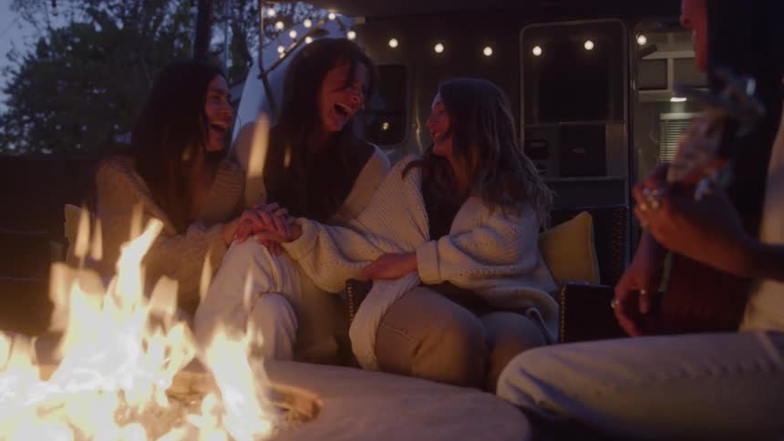 Four young female friends sitting beside a campfire outdoors next to an rv at sundown three seated close together talking and laughing while another sits across from them playing and singing with a guitar