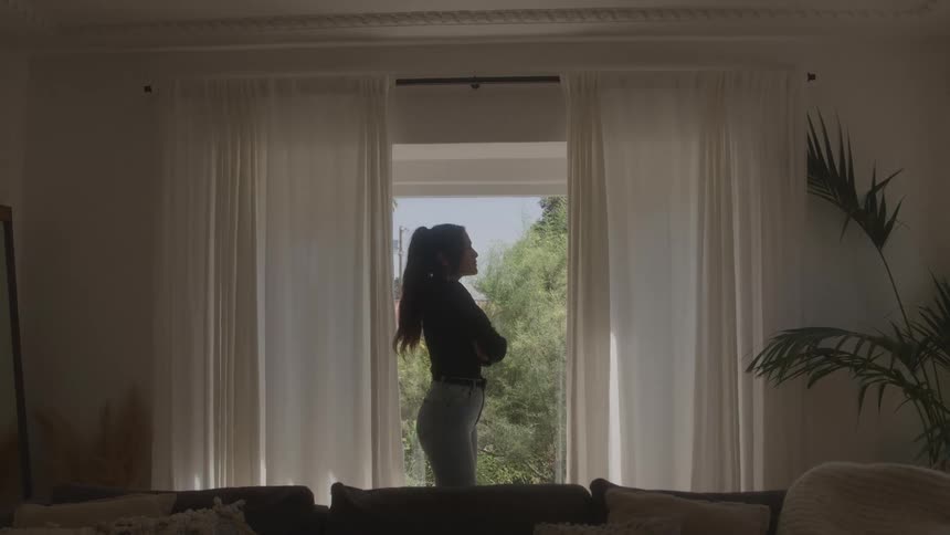 A side view of a woman in a black shirt and jeans standing in a living room as she looks out a large window with white curtains to a yard with green trees