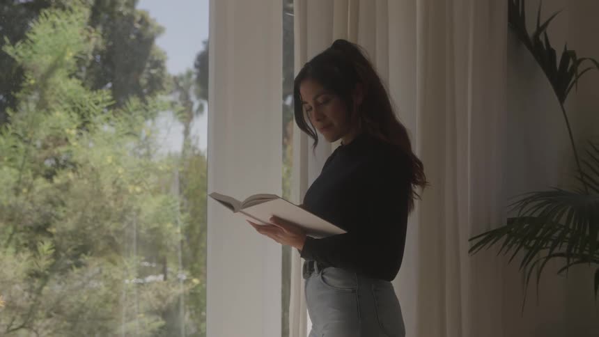 A woman in a black shirt and jeans stands by a window and reads a white book with a large tree visible outside