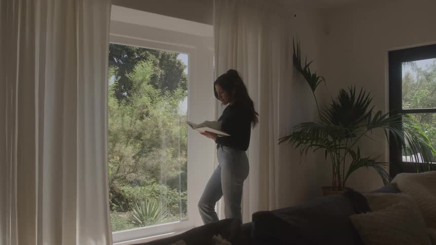 Young woman standing beside a window in her living room at midday reading a book with the curtains spread apart to reveal tree lines outside