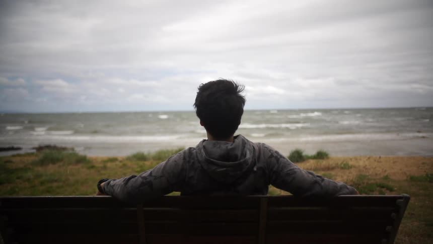 A man sitting on a bench near the ocean gazing out at the cloud-filled horizon as large waves roll towards shore