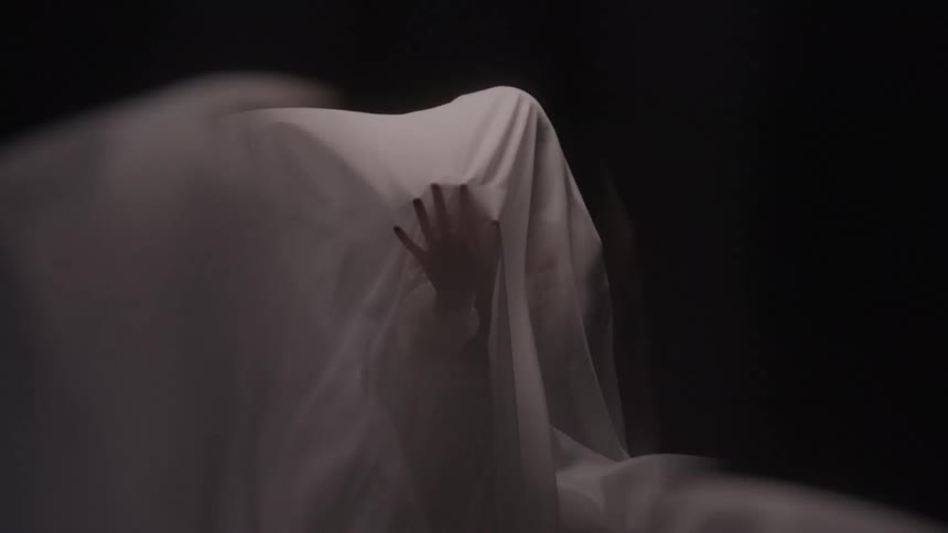 A persons head and shoulders are covered by a thin white sheet as they move and a hand presses outward from the inside