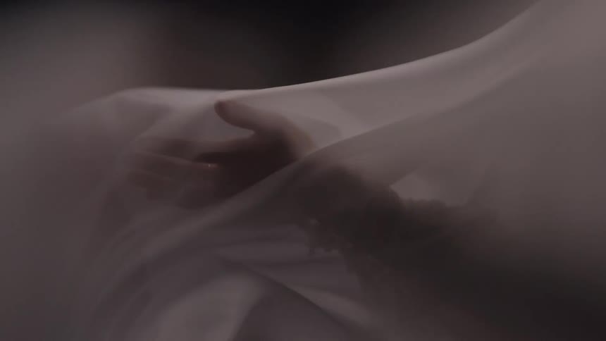 A close view shows a hand reaching out from underneath a thin white sheet that is draped over a persons body
