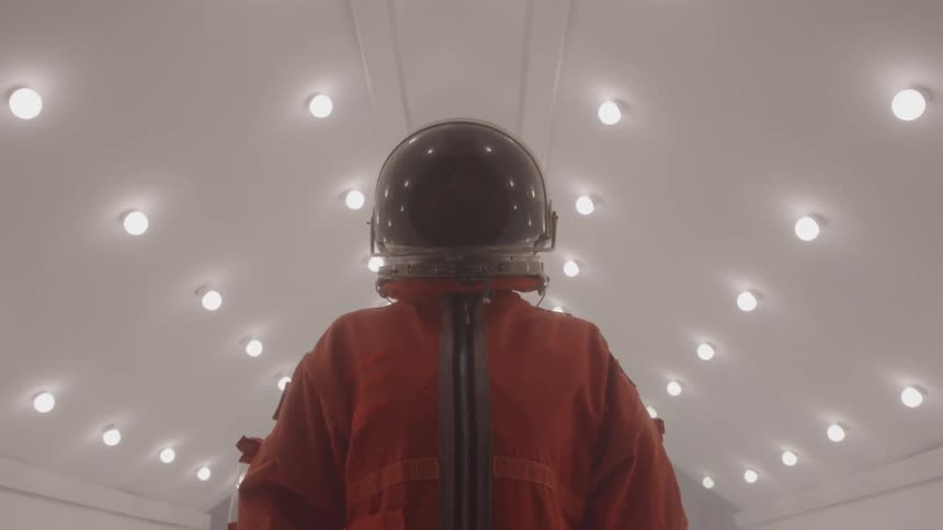 Person with helmet and orange spacesuit in a white lit hallway