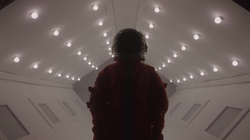 A person wearing a spacesuit is standing in a brightly lit corridor with a futuristic scifi aesthetic the person is silhouetted against the light facing away from the camera and looking ahead