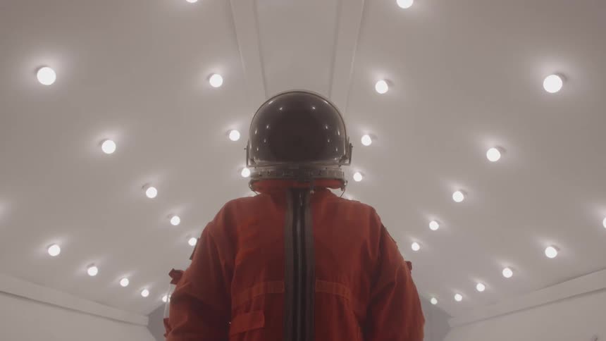 A person in a full orange space suit and a helmet stands on a sterile white corridor with many circular lights