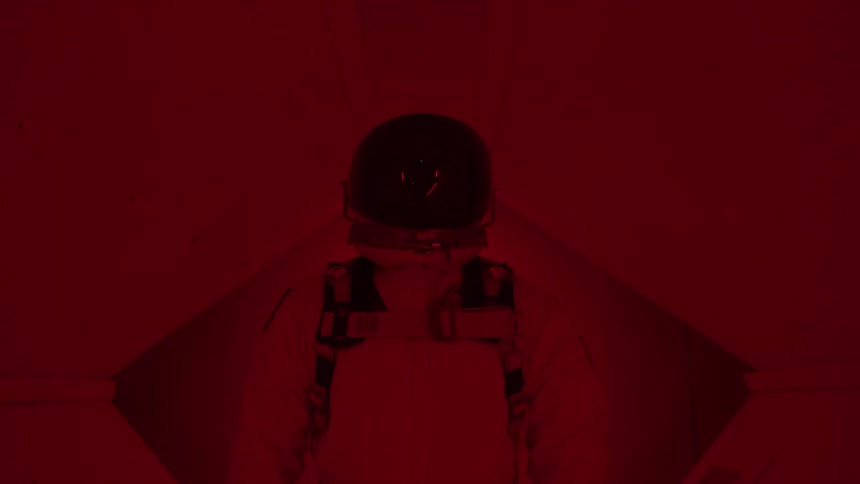 A wide view from behind shows a person in a white space suit and helmet standing in a sterile white room filled with a deep red and blue flashing lights