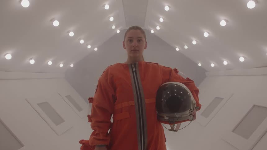 A woman in a bulky orange space suit stands looking at the camera while holding a dark helmet down by her side in a sterile white corridor with many circular lights on the ceiling