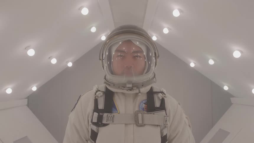 A man wearing a white and gray space suit and a helmet stands looking at the camera in a sterile white corridor with many circular lights