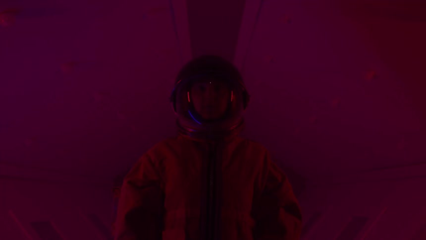 A person in an astronaut suit stands in a dark room with red lighting the person is looking directly at the camera with a determined expression appearing ready for their mission