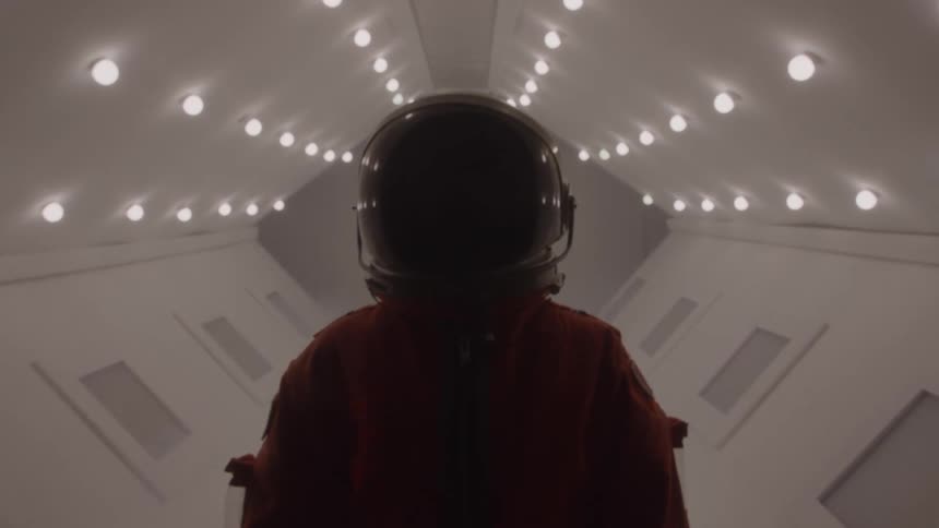 A person wearing an orange spacesuit and a helmet is standing in a brightly lit futuristic corridor the lighting creates a strong contrast against the persons silhouette as they look straight ahead