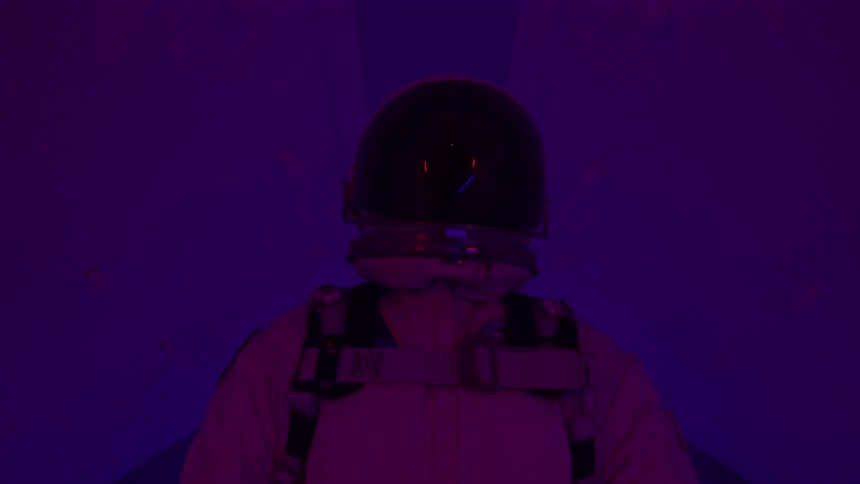 A wide view from behind shows a person in a white space suit and helmet standing in a sterile white room filled with a deep red and blue flashing lights
