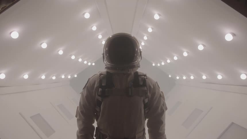 Person wearing a spacesuit and helmet inside a space chamber surrounded by a series of lights