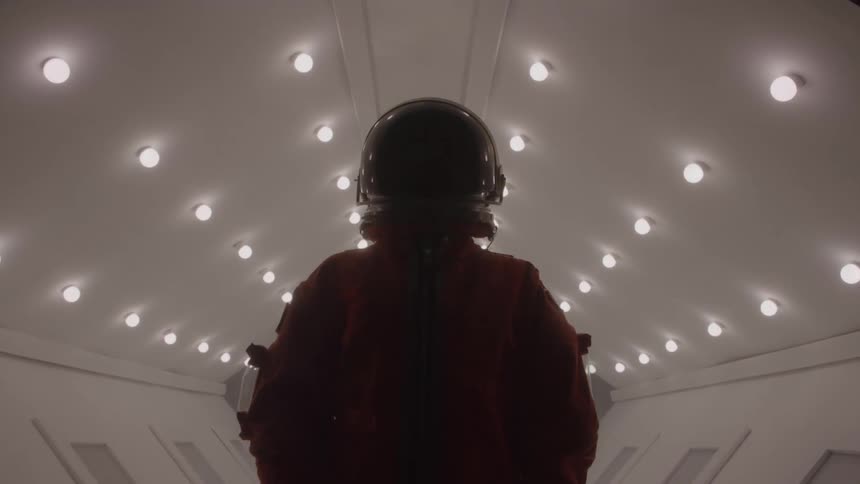 A person wearing a spacesuit and a helmet is standing in a brightly lit futuristic corridor the lighting creates a strong contrast silhouetting the person as they stand facing away from the camera